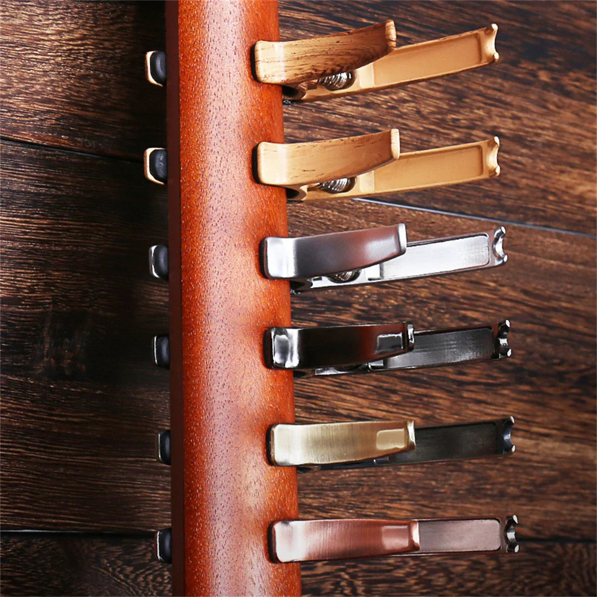 A wooden rack holding six guitar capo metal clips with zinc alloy construction in gold silver and black finishes