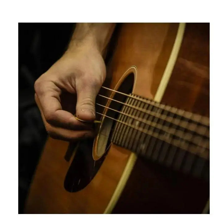 A hand holding a golden guitar pick near the strings of a warm brown acoustic guitar from janyun 150 pcs guitar picks sampler