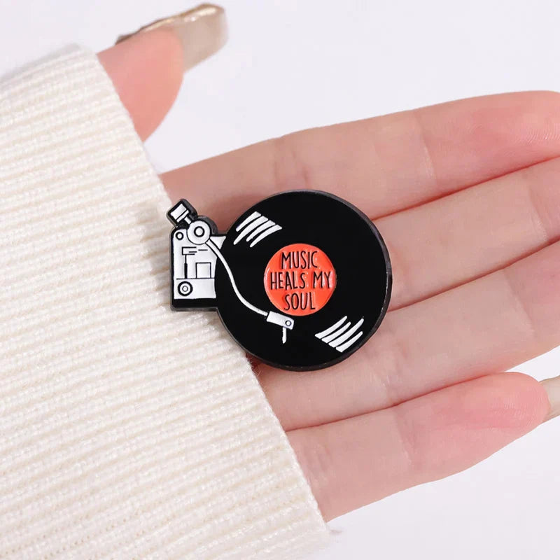 Black enamel pin shaped like vinyl record with white grooves orange center and music heals my soul text for 90s retro music fans