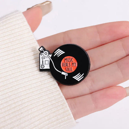 Black enamel pin shaped like vinyl record with white grooves orange center and music heals my soul text for 90s retro music fans