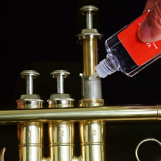 A hand applies clear valve oil from a red-labeled bottle to the brass instrument valve for music instrument lubrication