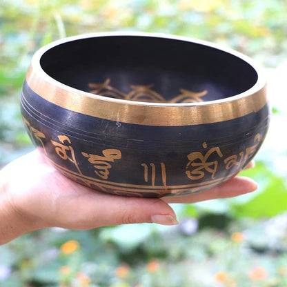 A hand holds a black metal singing bowl with gold rims and Sanskrit inscriptions for bell yoga meditation