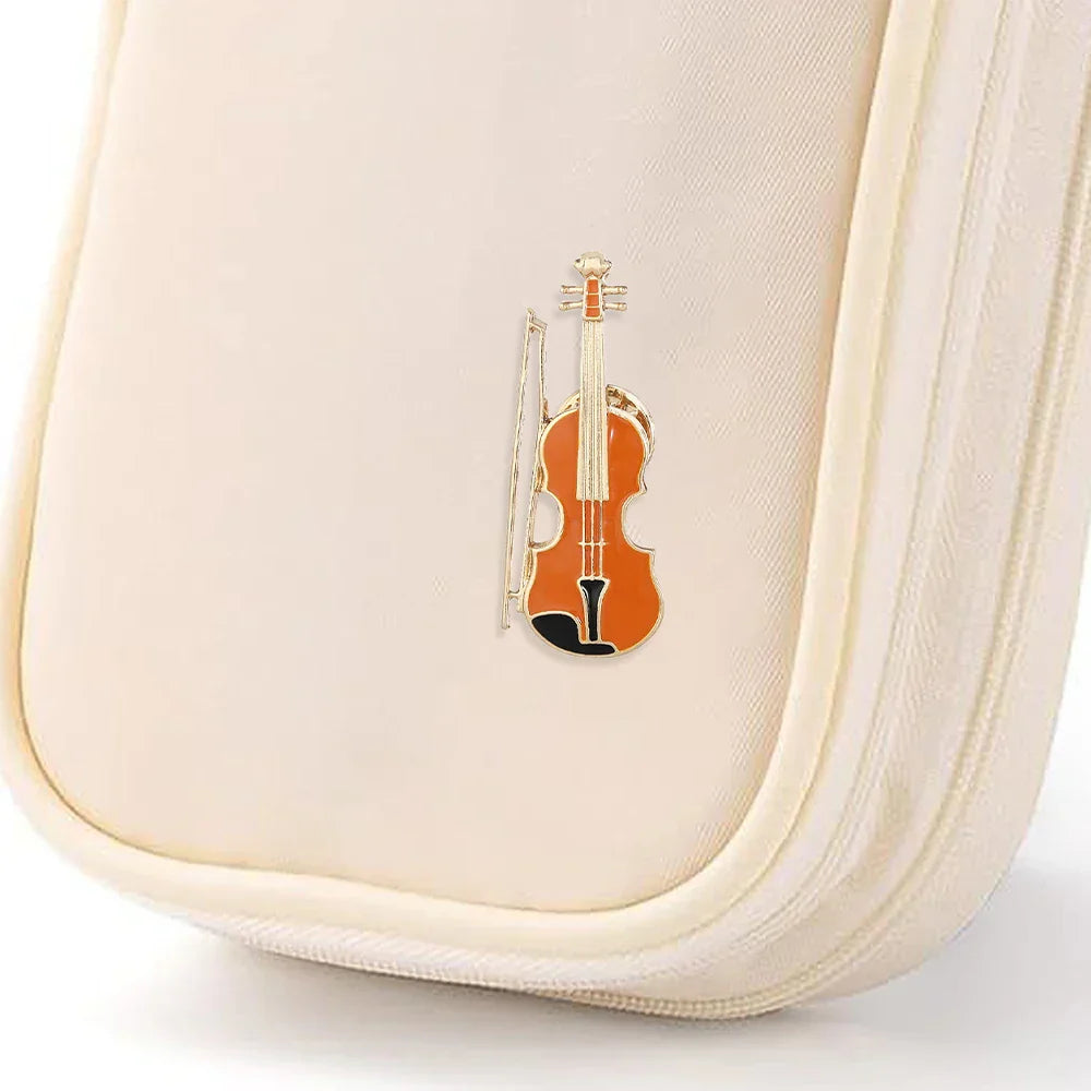 A small orange and black enamel violin pin with a silver bow attached to an off-white pouch from instruments enamel brooches collection