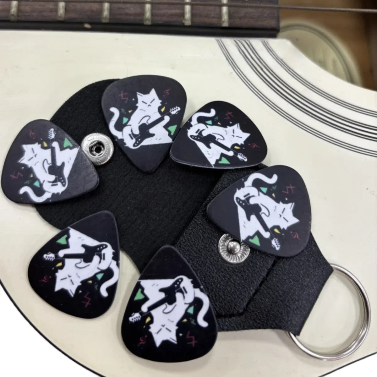 Black guitar picks double-sided printed with white cartoon cats in green and red details held in black leather keychain holster