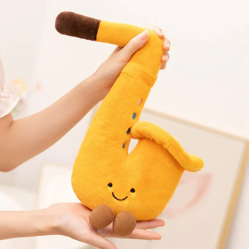 Yellow plush saxophone with brown mouthpiece and feet featuring embroidered eyes and smile from musical instrument series plush toy