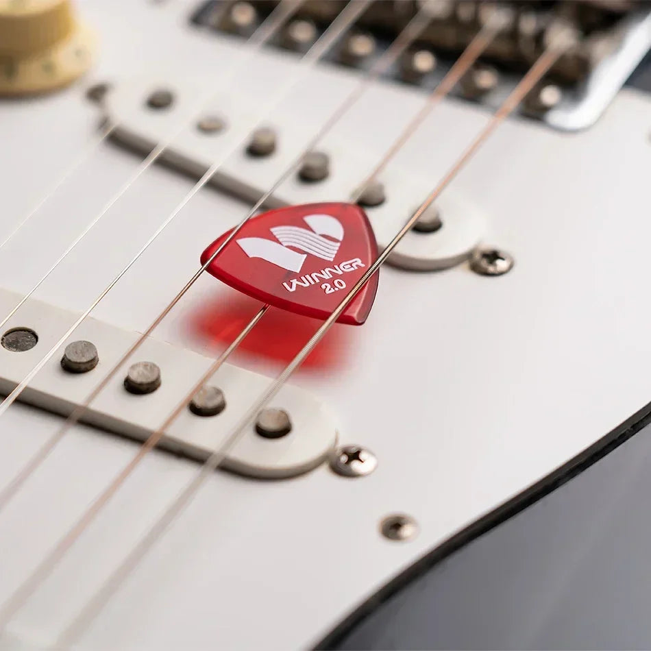 Red triangular winner hybrid polycarbon guitar pick 2.0mm resting on white acoustic electric guitar strings with box guitar accessories
