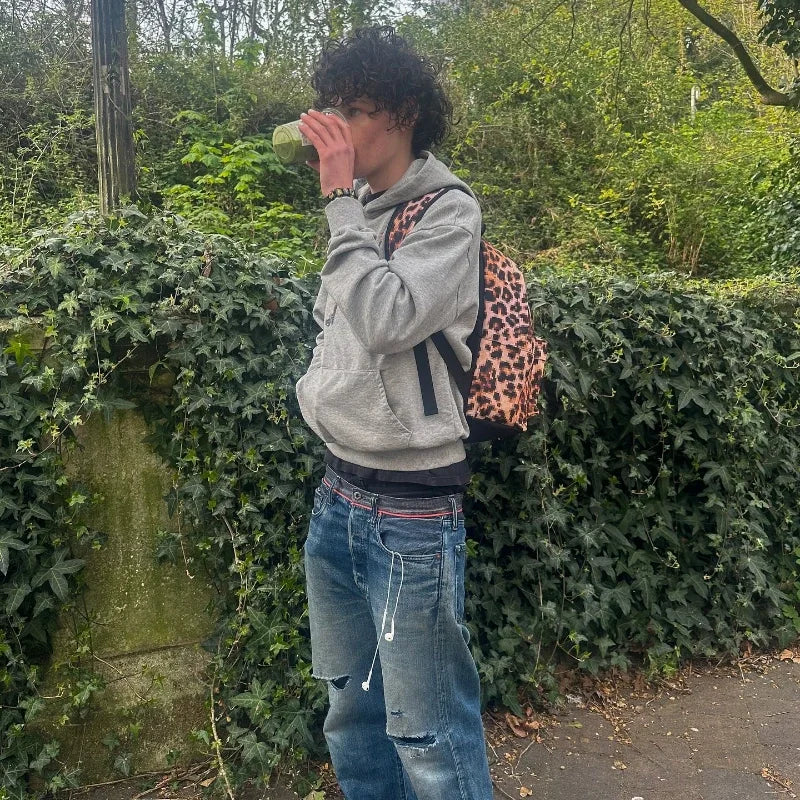 A person wearing a y2k gray sports hoodie retro smoke print sweater with ripped blue jeans and a leopard print backpack